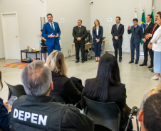 Nesta foto, o Defensor Público-Geral discursa de frente para a câmera. Em pé, logo atrás dele, um grupo de autoridades acompanha o pronunciamento. Em primeiro plano, vemos a plateia sentada de costas, assistindo à cerimônia. Um detalhe notável ao fundo, no canto esquerdo, é um pequeno espaço infantil com tapete colorido e brinquedos, sugerindo o caráter acolhedor do novo local.