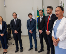 Em primeiro plano, no lado direito da imagem, a Ouvidora-Geral da DPE-PR faz seu discurso. Ela tem uma expressão séria e mantém as mãos unidas à frente do corpo. Ao seu lado, um grupo de autoridades, composto por quatro homens e uma mulher, ouve atentamente. Ao fundo, as bandeiras do Brasil e do Paraná estão hasteadas, compondo o cenário oficial.