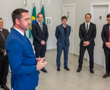 A foto foca no Defensor Público-Geral durante seu discurso, visto em primeiro plano. Com o rosto concentrado, ele gesticula e se dirige a um grupo de cinco homens que o ouvem atentamente. Os homens estão em pé, formando um semicírculo. O ambiente é uma sala ampla com paredes claras e boa iluminação.