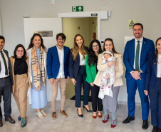 Esta é uma foto de confraternização, que reúne um grupo posando para a câmera. Estão presentes a Ouvidora-Geral, defensores da sede de Foz, o Defensor Público-Geral e a Primeira e Segunda Subdefensoras-Gerais. São dez pessoas no total, além de um bebê no colo de uma das participantes. Todos sorriem em um corredor do novo prédio, onde uma placa na parede identifica o local: "DEFENSORIA PÚBLICA DO ESTADO DO PARANÁ".