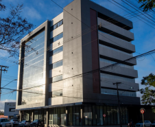 Esta foto exibe edifício da sede atual da DPE-PR em Foz do Iguaçu. Trata-se de um prédio grande e moderno de múltiplos andares. O céu está azul e ensolarado. A fachada do prédio é uma combinação de vidro espelhado, que reflete o céu, e painéis de cor cinza-escuro. A arquitetura tem linhas retas e um design contemporâneo.