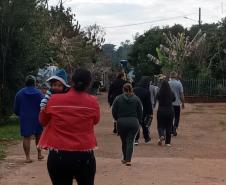 Imagem mostra um grupo de pessoas caminhando por rua de barro. No primeiro plano uma mulher carrega um bebê. 