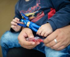 Imagem mostra em close uma criança sentada no colo do pai segurando um boneco de super herói enquanto a aguarda atendimento no Mutirão Meu Pai Tem Nome. 