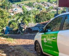 Imagem mostra carro da DPE-PR em rua calçada com pedras e ao fundo casas em meio à plantações. 