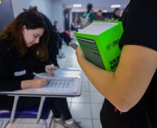 Imagem mostra braço de candidata segurando livro em close no primeiro plano enquanto fiscal confere seus documentos para ingressar na sala para do concurso. 