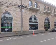 Imagem que mostra o prédio da nova sede da DPE em União da Vitória. O prédio possui fachada do andar térreo revestida em pedras de cor clara e marcada por grandes janelas e uma porta em formato de arco ogival. O andar superior também apresenta janelas em arco com sacadas e grades de ferro. À esquerda, uma porta de vidro exibe o logotipo e o nome da instituição. Através do vidro, é possível ver parte do interior da recepção.