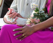 Uma imagem focada nas mãos de um casal. A noiva, usando um vestido vibrante de cor magenta, tem uma aliança dourada no dedo e as unhas pintadas. Sua mão repousa sobre a perna, próxima a um buquê. As mãos do noivo, que veste uma camisa clara, estão unidas e adornadas com um pequeno arranjo de margaridinhas amarrado em seu pulso.