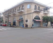 Imagem que mostra o prédio da nova sede da DPE em União da Vitória visto a partir de uma esquina. Há semáforos para carros e pedestres, e várias pessoas estão caminhando e atravessando a rua. Um grupo de bicicletas está estacionado na calçada. Carros estão parados na rua à esquerda e uma motocicleta passa pela direita. O céu está encoberto, indicando um dia nublado.
