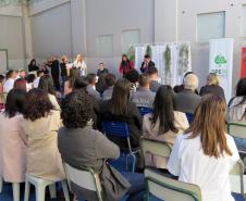 Esta foto oferece uma visão mais ampla da cerimônia, tirada de trás dos convidados. A plateia está sentada em cadeiras simples, observando os casais e o celebrante no palco. No lado direito, são visíveis dois banners: um do "Conselho da Comunidade de Londrina" e outro da Defensoria Pública do Estado do Paraná.