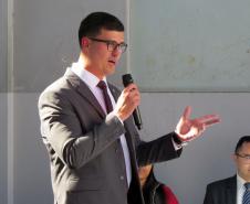 Imagem do defensor Pedro Bruzzi discursando com um microfone na mão. Ele veste um terno cinza e óculos, sua expressão e gestos indicam que ele está fazendo um pronunciamento como uma das autoridades do evento. Ao fundo, outra pessoa pode ser vista sentada.