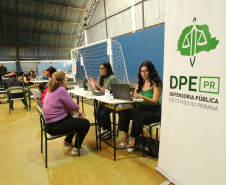 Imagem que mostra um ginásio de esportes adaptado para o atendimento. Uma mulher de blusa roxa é acolhida por duas integrantes da equipe da Defensoria Pública do Paraná. Enquanto uma das atendentes dialoga e gesticula, a outra opera um notebook. Há um banner da DPE-PR, em primeiro plano à direita.
