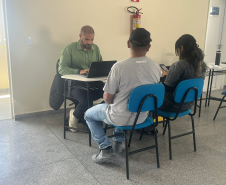 Imagem que mostra uma cena de atendimento individual. Três pessoas estão agrupadas em torno de uma pequena mesa branca. À esquerda, de frente para a mesa, está o atendente. Ele é um homem de pele morena, com cabelo escuro bem curto e barba. Veste uma camisa social de manga longa e está concentrado. Do outro lado da mesa, de costas para a câmera, estão duas pessoas sentadas em cadeiras com assentos e encostos azuis, um homem e uma mulher.