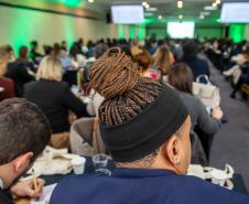 Homem negro com tranças e homem branco tomando notas em caderno, ambos de costas em primeiro plano, e ao fundo se vê a sala lotada de defensores de too o país na abertura di Conajuri.