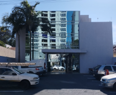 Imagem que mostra a fachada da sede da Defensoria Pública em Cianorte, em um dia claro e ensolarado. O prédio é uma construção moderna de dois andares, com uma grande seção central de vidro espelhado. As laterais do prédio são de alvenaria, pintadas em um tom de cinza-claro. Na frente do edifício há um estacionamento com alguns carros parados. À esquerda do prédio, uma palmeira alta se destaca contra o céu.