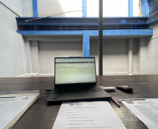A imagem é uma visão em primeiro plano de uma mesa de trabalho de madeira. Sobre a mesa, há um laptop aberto e formulários de questionário para entrevistas. O ambiente é o pátio interno da cadeia, com paredes de concreto e estruturas pintadas de azul na parte superior.