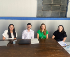 Quatro pessoas, três mulheres e um homem, posam para uma foto, sentados lado a lado atrás de uma mesa de madeira. Eles estão em um local com parede branca e uma faixa azul, com uma janela gradeada ao fundo.