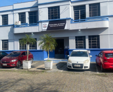 A foto mostra a fachada da Cadeia Pública de Paranaguá, um prédio de dois andares, pintado em tons de azul claro e escuro, sob um céu limpo. Em frente, há um estacionamento com carros.