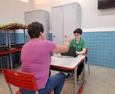 Em uma sala de aula, uma mulher de blusa rosa está sentada de costas para a câmera, em frente a uma mesa. Ela entrega um documento para uma servidora da Defensoria, uma jovem de camisa verde e óculos que está do outro lado da mesa, sorrindo e usando um laptop.
