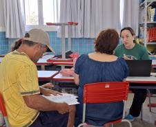 Dentro de uma sala de aula, um casal é atendido por uma servidora da Defensoria Pública de camisa verde. O homem, de camiseta amarela e boné, lê alguns papéis, enquanto a mulher, de blusa jeans e de costas para a foto, ouve a explicação da servidora.