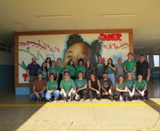 Uma foto de grupo com cerca de vinte pessoas, a equipe da Defensoria Pública, do CRAS Rural e da Escola Municipal posando em um pátio coberto de uma escola. Eles estão em pé e agachados em frente a um grande mural colorido pintado na parede ao fundo.