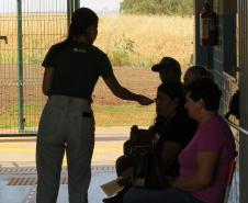 Vista de dentro de um pátio, uma servidora está em pé, conversando com um grupo de três pessoas sentadas em um banco. Ao fundo, através de uma grade, vê-se um campo aberto com vegetação seca sob a luz do sol.