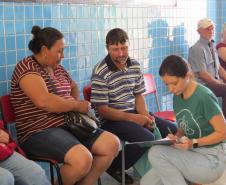 Em um corredor com parede de azulejos azuis, pessoas aguardam sentadas. Em primeiro plano, uma servidora de camisa verde está agachada, anotando informações em uma prancheta enquanto conversa com um casal sentado em cadeiras vermelhas.