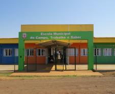 A foto mostra a fachada colorida de uma escola rural, com paredes pintadas em azul, laranja e verde. Um grande portal verde sobre a entrada tem a inscrição "Escola Municipal do Campo, Trabalho e Saber". O dia está ensolarado, com céu azul e sem nuvens.
