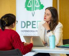 Imagem que mostra, em uma mesa de atendimento, uma defensora pública de blazer branco que escuta uma mulher. A pessoa atendida está de costas e usa um suéter vermelho vivo. A defensora, com a mão no queixo, olha atentamente para a mulher, com um laptop aberto entre elas. Ao fundo, um banner da Defensoria Pública é visível.