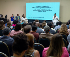Um auditório cheio com a plateia sentada de costas para a foto, voltada para um palco. No palco, uma mulher de branco fala ao microfone ao lado de uma tela projetando o título de um curso sobre heteroidentificação racial.