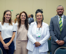 Quatro pessoas posam lado a lado para uma foto em um ambiente interno. Da esquerda para a direita: uma mulher de blusa branca, uma mulher de roupa bege, uma mulher negra com tranças e roupa branca, e um homem negro de terno cinza e gravata verde.