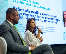 Uma mulher de cabelos longos e blusa branca fala ao microfone, sentada ao lado de um homem de terno. Atrás deles, um telão exibe o texto "Ações afirmativas por meio de cotas raciais e heteroidentificação".