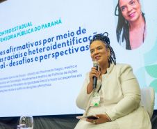 Uma mulher negra com tranças e roupa branca sorri enquanto fala ao microfone, sentada no palco. Ao fundo, uma tela projeta sua foto, nome e o tema da palestra sobre ações afirmativas e cotas raciais.