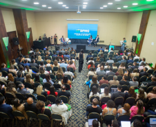 Vista superior do fundo de um auditório lotado, com a plateia voltada para um palco com iluminação azul. No telão, lê-se "Modelos de Reparação Coletiva da DPMG", enquanto palestrantes debatem sentados no palco.