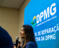 Uma mulher de perfil, com cabelos longos, fala ao microfone. Atrás dela, um painel de LED azul exibe o logotipo da DPMG e o texto parcial "Modelos de Reparação Coletiva".