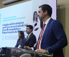 Um homem de terno azul e gravata vermelha discursa em um púlpito. Atrás dele, duas pessoas estão sentadas no palco, e um telão exibe o título da palestra: "Litigância Estratégica com foco no extrajudicial".