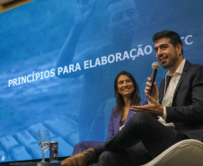 Um homem de terno escuro e camisa branca fala ao microfone, sentado em uma poltrona branca no palco. Ao seu lado, uma mulher de blazer azul sorri enquanto observa a plateia. Atrás deles, um grande painel de LED azul exibe a frase "PRINCÍPIOS PARA ELABORAÇÃO".