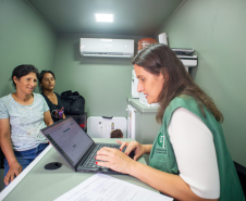 Defensora Mariana, com colete verde da Defensoria, atende duas mulheres, uma com blusa azul claro e outra com camiseta escura. Elas estão no interior da van dos direitos. Ao fundo um ar condicionado e uma mesinha com impressora.