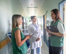 Defensora Ingrid e defensora Mariana, conversando com funcionária de hospital local. Elas estão em um corredor com paredes claras, ao fundo uma luz de contraste com funcionária.