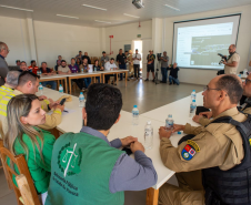 Cena de reunião com várias pessoas sentadas em mesas brancas em "U" em uma sala iluminada. Em primeiro plano, defensores públicos juntos de oficiais da Polícia Militar e outras autoridades, sentados. Há uma tela de projeção exibindo um mapa ao fundo.