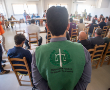 Cena de reunião com várias pessoas sentadas em mesas brancas em "U" em uma sala iluminada. Em primeiro plano, defensores públicos juntos de oficiais da Polícia Militar e outras autoridades, sentados. Há uma tela de projeção exibindo um mapa ao fundo.