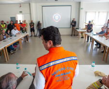 Em uma sala de reunião, o secretário das cidades do Estado, Guto Silva, está em pé vestindo colete laranja (Defesa Civil/Casa Militar), fala com o público sentado nas mesas brancas.