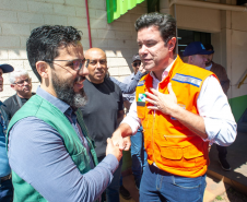 Dois homens brancos se cumprimentam com um aperto de mãos. O da esquerda, o defensor público Fernando Redede, com barba e óculos, veste um colete verde da defensoria. À direita, o secretário das cidades do Estado, Guto Silva, veste um colete de segurança laranja, com a mão esquerda sobre o peito. Há outras pessoas ao fundo.