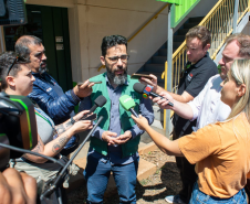 O defensor público, Fernando Redede - um homem de barba e colete verde da Defensoria Pública - é cercado por repórteres que seguram microfones com logotipos de emissoras, em uma entrevista rápida ao ar livre.