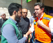 Dois homens brancos se cumprimentam com um aperto de mãos. O da esquerda, o defensor público Fernando Redede, com barba e óculos, veste um colete verde da defensoria. À direita, o secretário das cidades do Estado, Guto Silva, veste um colete de segurança laranja, com a mão esquerda sobre o peito. Há outras pessoas ao fundo.