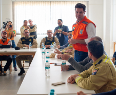 Em uma sala de reunião, o secretário das cidades do Estado, Guto Silva, está em pé vestindo colete laranja (Defesa Civil/Casa Militar), fala com o público sentado nas mesas brancas.