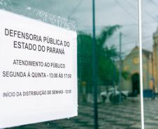 Imagem da placa da Defensoria com reflexo na porta de vidro da Câmara em que se vê o reflexo da igreja na praça. da igreja da praça em Matinhos.