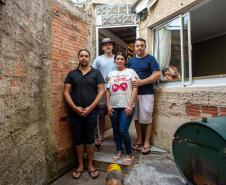 Família com quatro pessoas adultas posa em frente à sua casa. Muros e paredes sem reboco compõe o cenário.