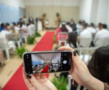 Imagem mostra um celular em primeiro plano e ao fundo a cerimônia com 20 casais. 