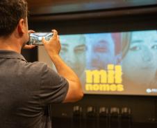 Um homem de costas fotografa com celular a tela com cartaz do documentário Mil Nomes.