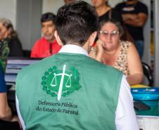 Imagem mostra Defensor Público de costas com colete da instituição realizando atendimento de uma senhora em mutirão no Litoral do Paraná.